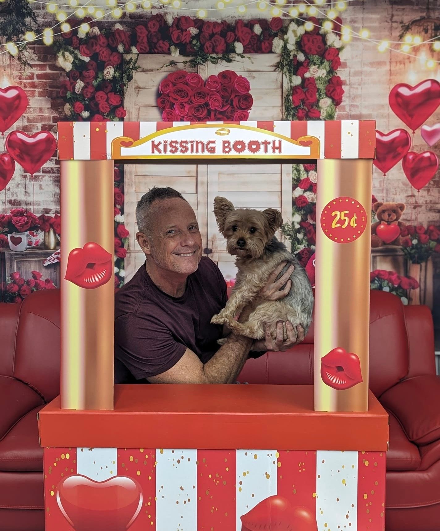 Man holding a small dog in a kissing booth, surrounded by Valentine's Day decorations.