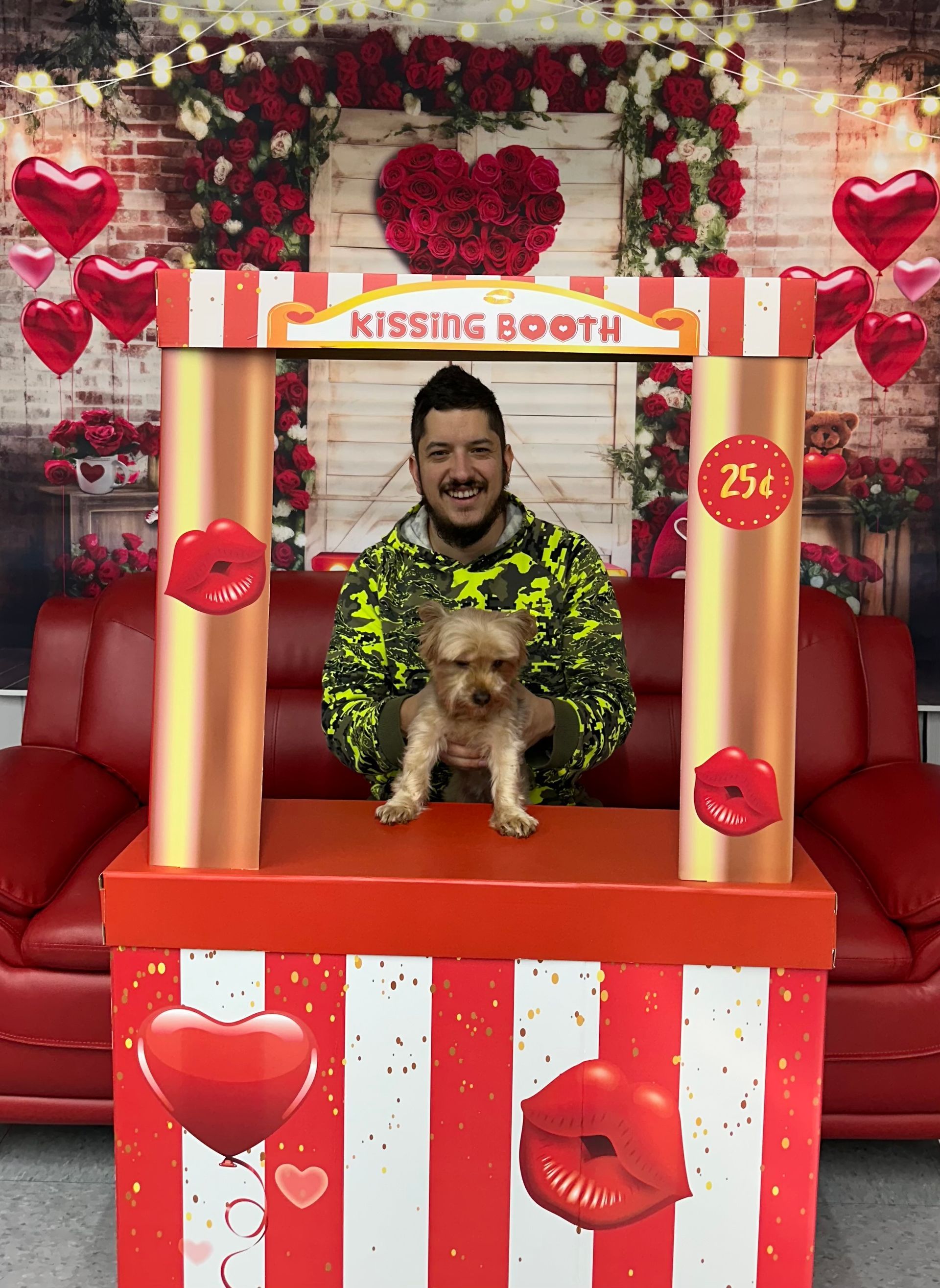 Man and small dog in Valentine's kissing booth; red, white, gold decor; smiling.