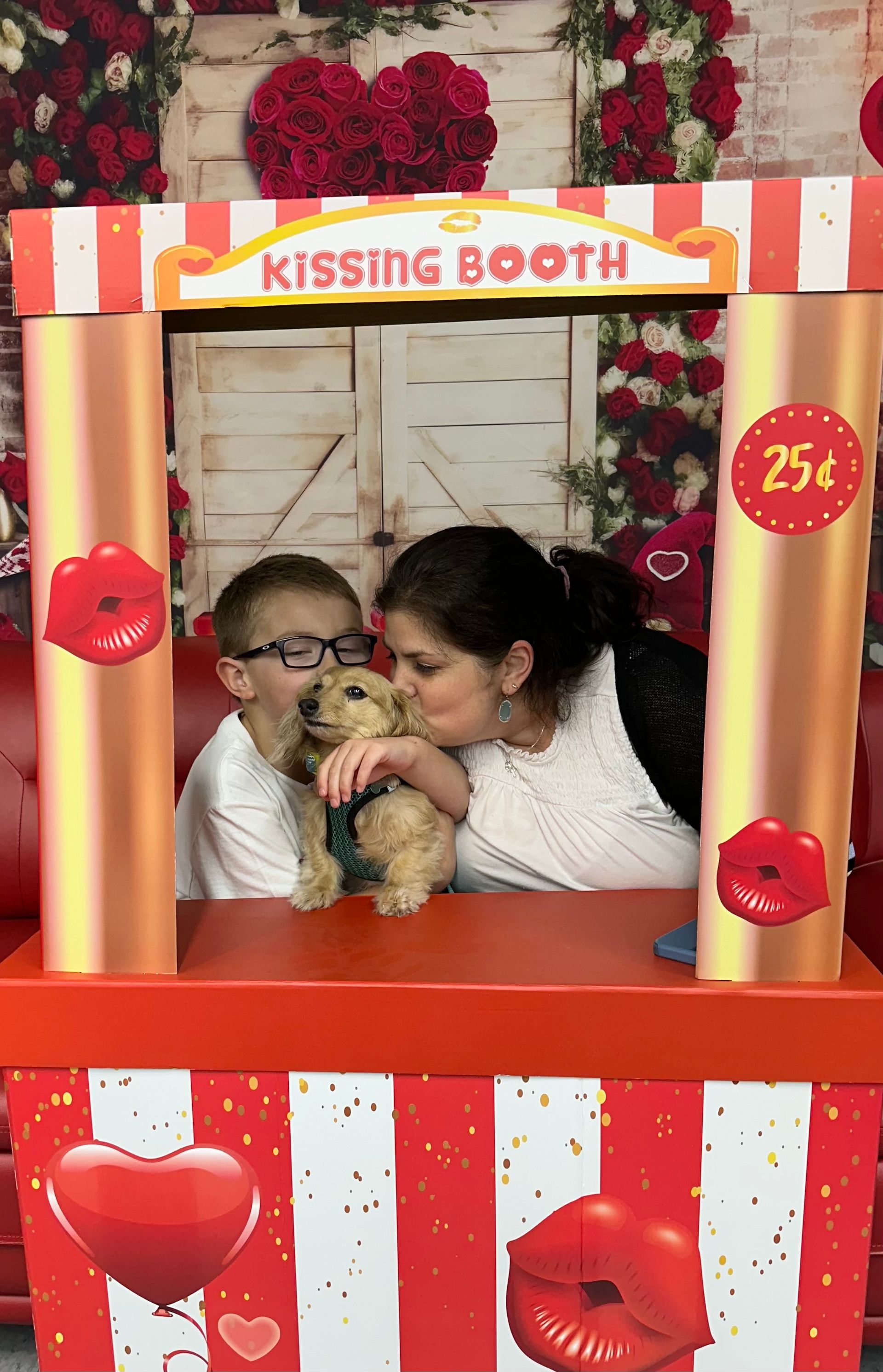 Boy, woman, and puppy in kissing booth; woman kissing puppy, boy smiling. Valentine's Day theme.