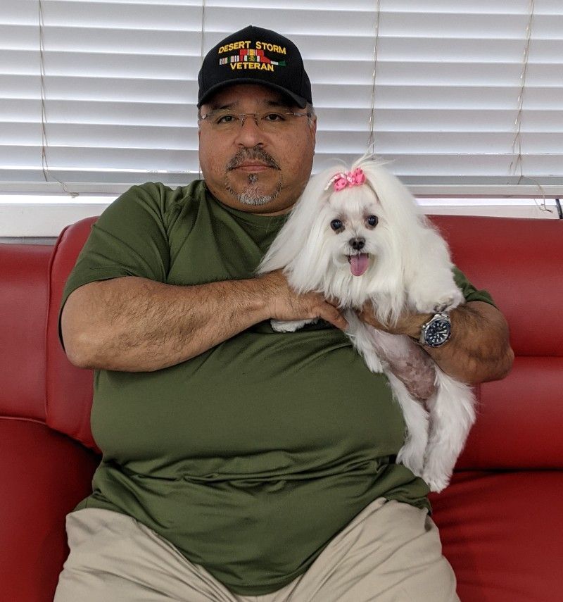 Man in cap holds small white dog with pink bow; both seated on red couch.