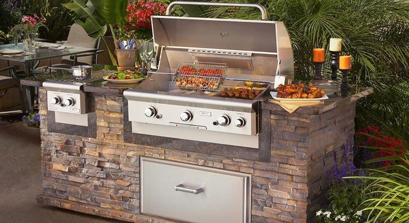 Outdoor stone barbecue island with stainless steel grill, side burner, and trays of food and drinks