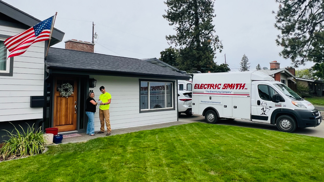 An electric service truck is parked by a house; two workers stand by the front door with an American flag nearby.