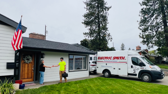 Residential service van parked outside a house; worker in neon shirt stands by the front door and an American flag hangs nearby.