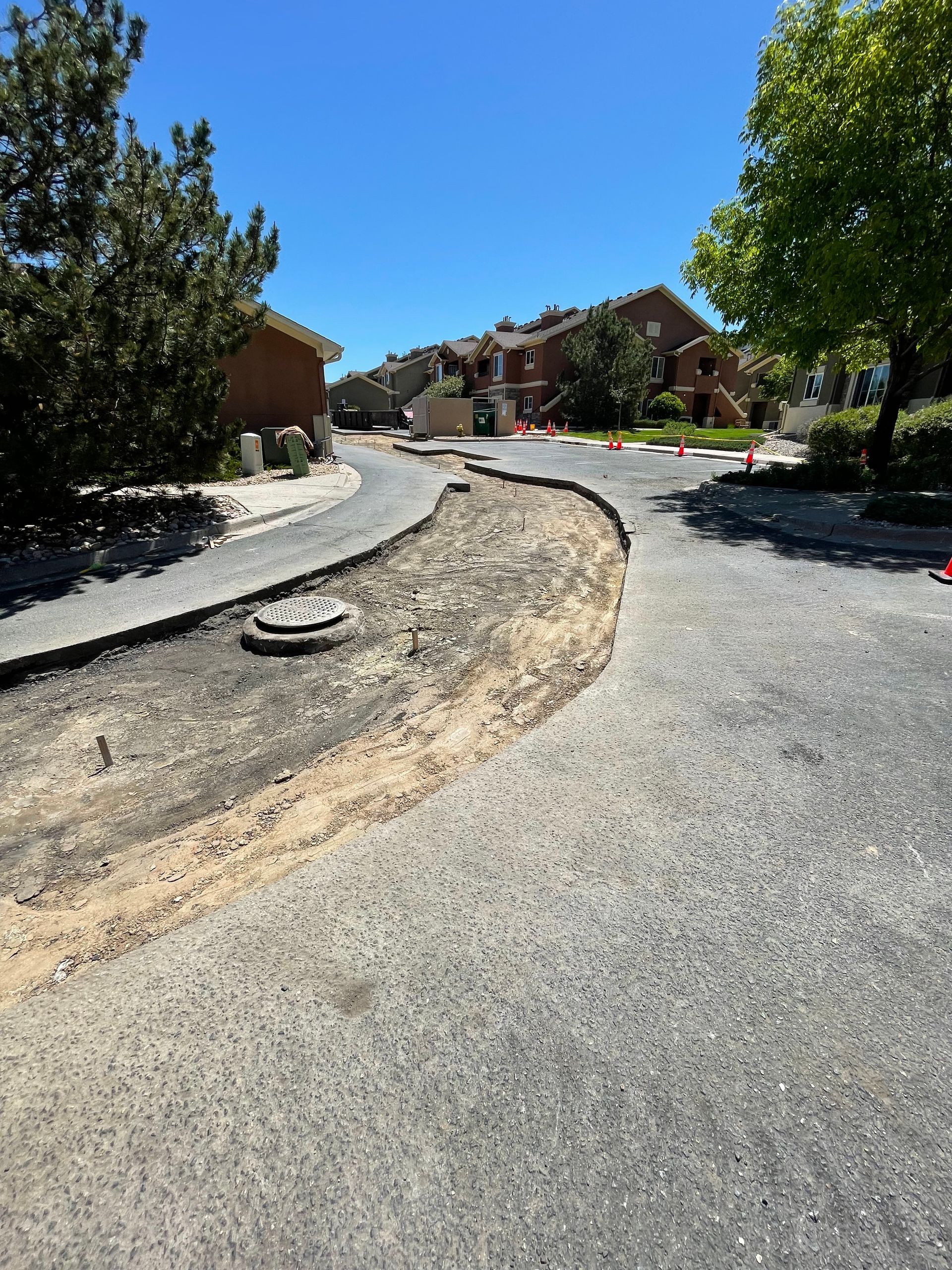 Asphalt path under repair with a bare dirt section, next to a curved path, residential buildings in the background, sunny day.