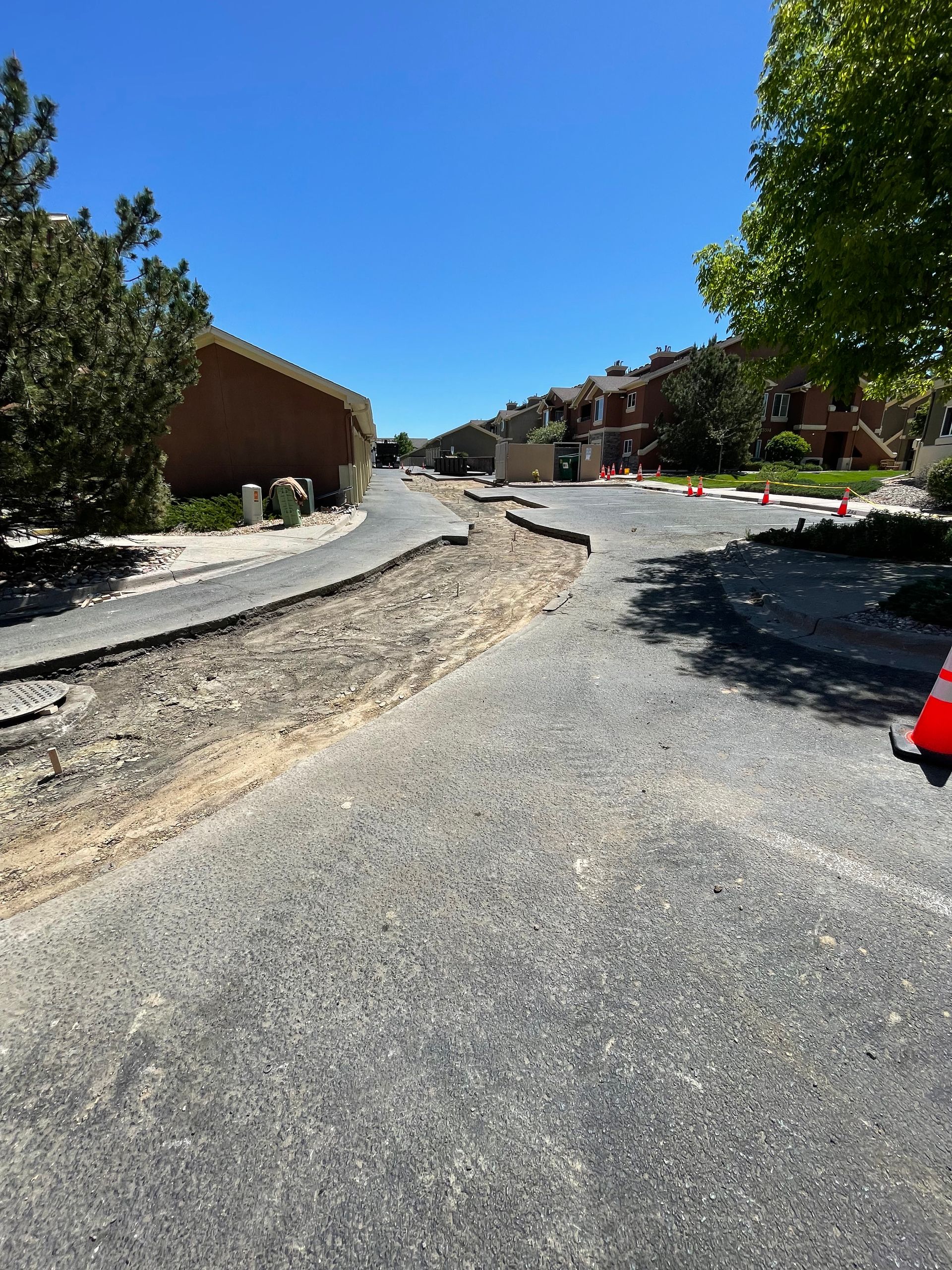 Asphalt road under construction.  Bare dirt and removed asphalt on the left side. Buildings and trees in the background under a clear blue sky.