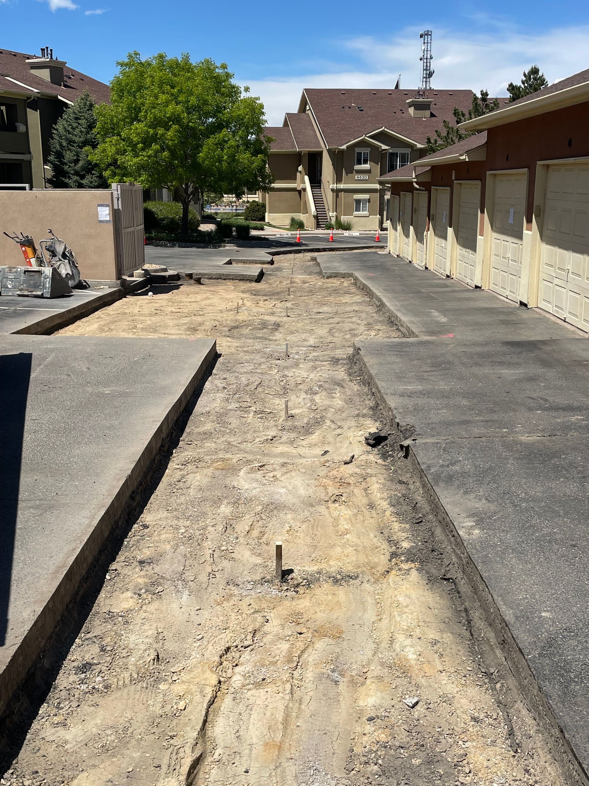 Driveway undergoing construction with concrete removed, leaving dirt and gravel. Beige apartment buildings and garages line the sides.