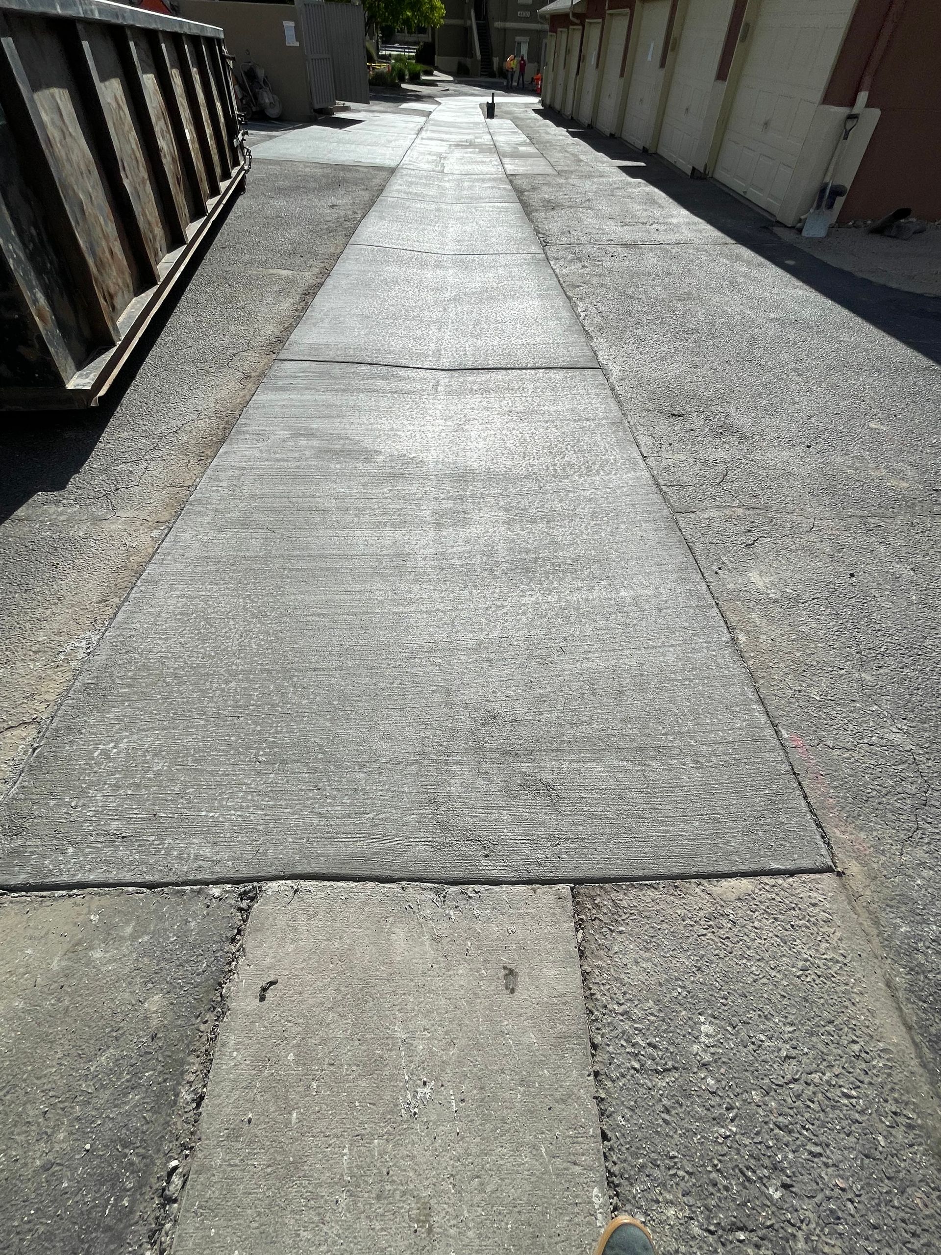 Concrete alleyway with a central strip and gravel on either side. A dumpster is on the left, and a building is on the right.