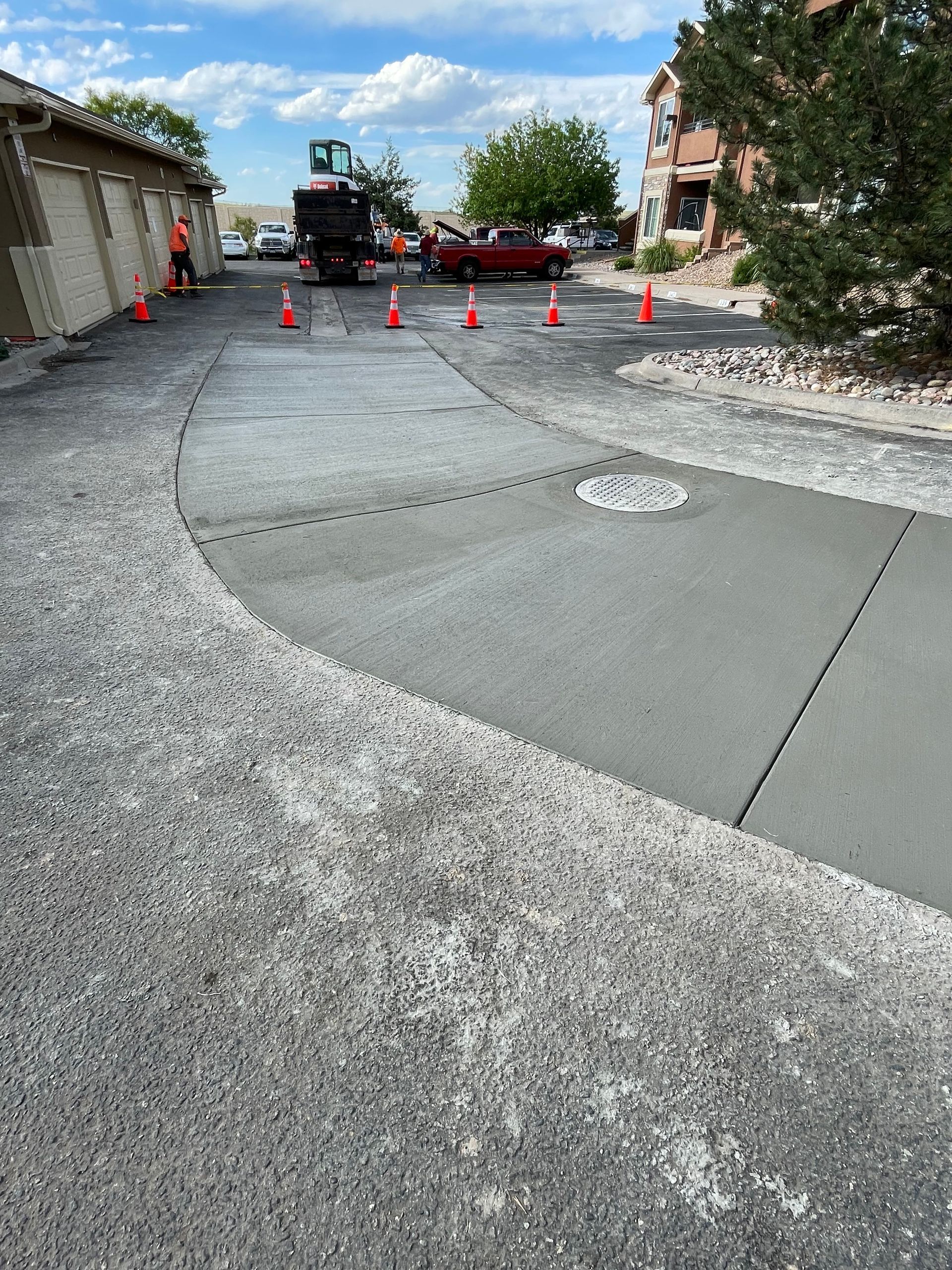 Freshly poured concrete driveway section with construction cones and machinery, adjacent to a gravel path, garages, and a residential building.