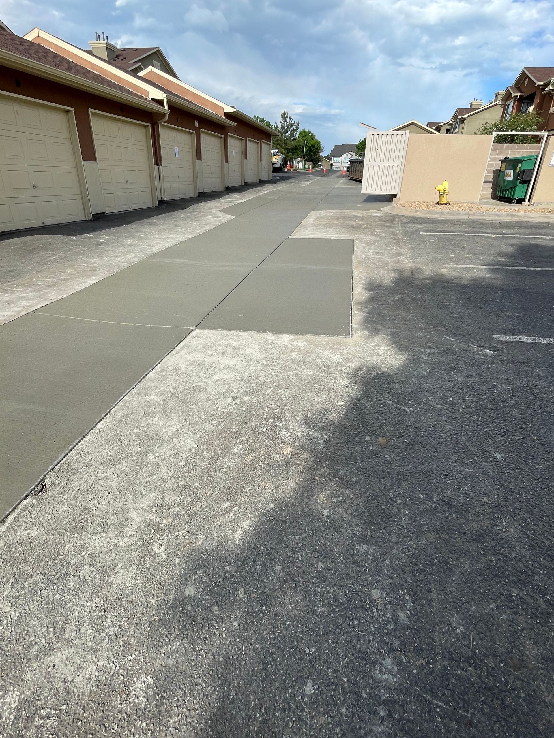 Asphalt driveway with patched areas, between attached beige garages with brown trim. Cloudy sky overhead.