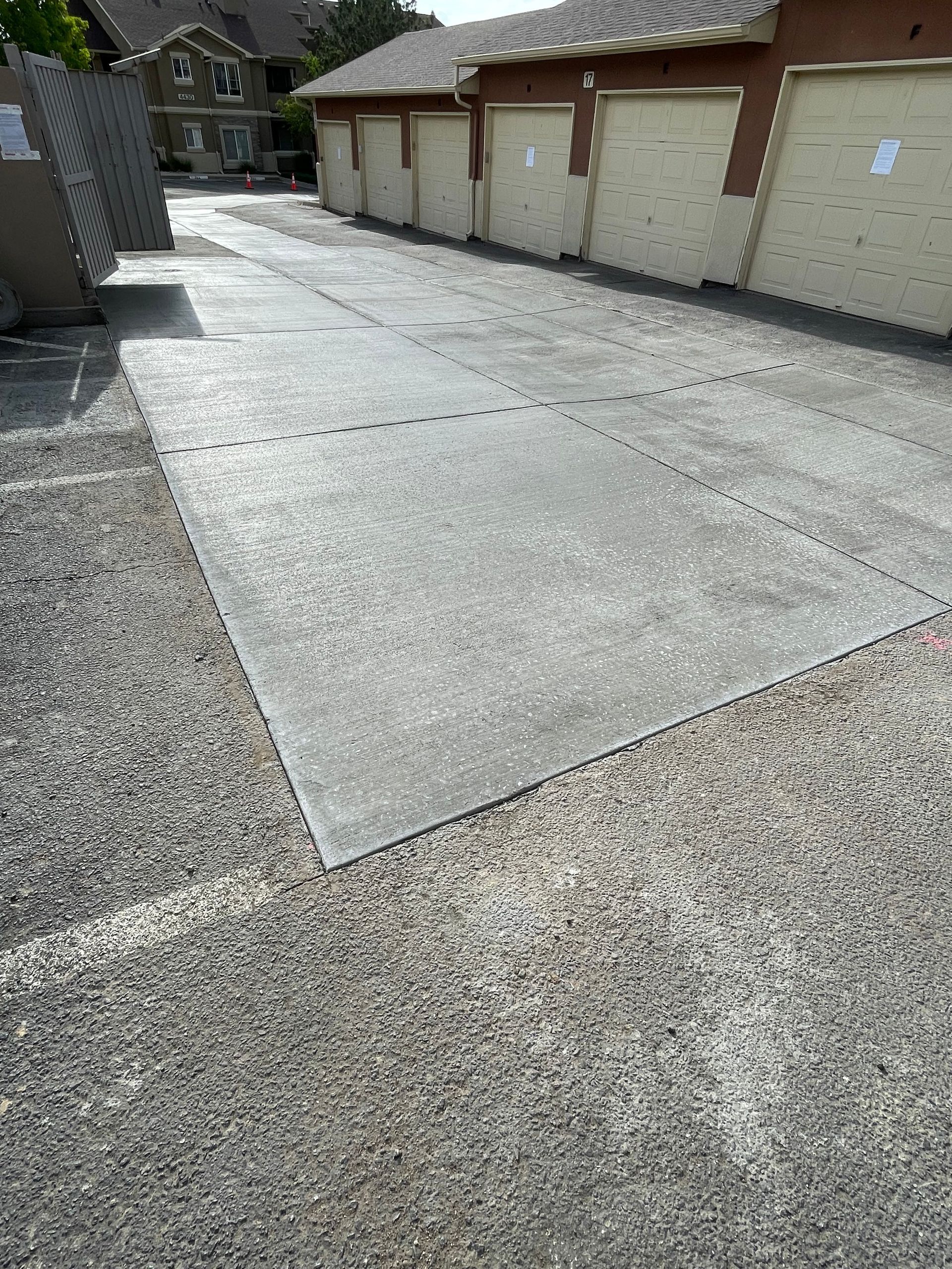 Asphalt parking area next to a row of tan garage doors, with a gravel border and a building in the background.