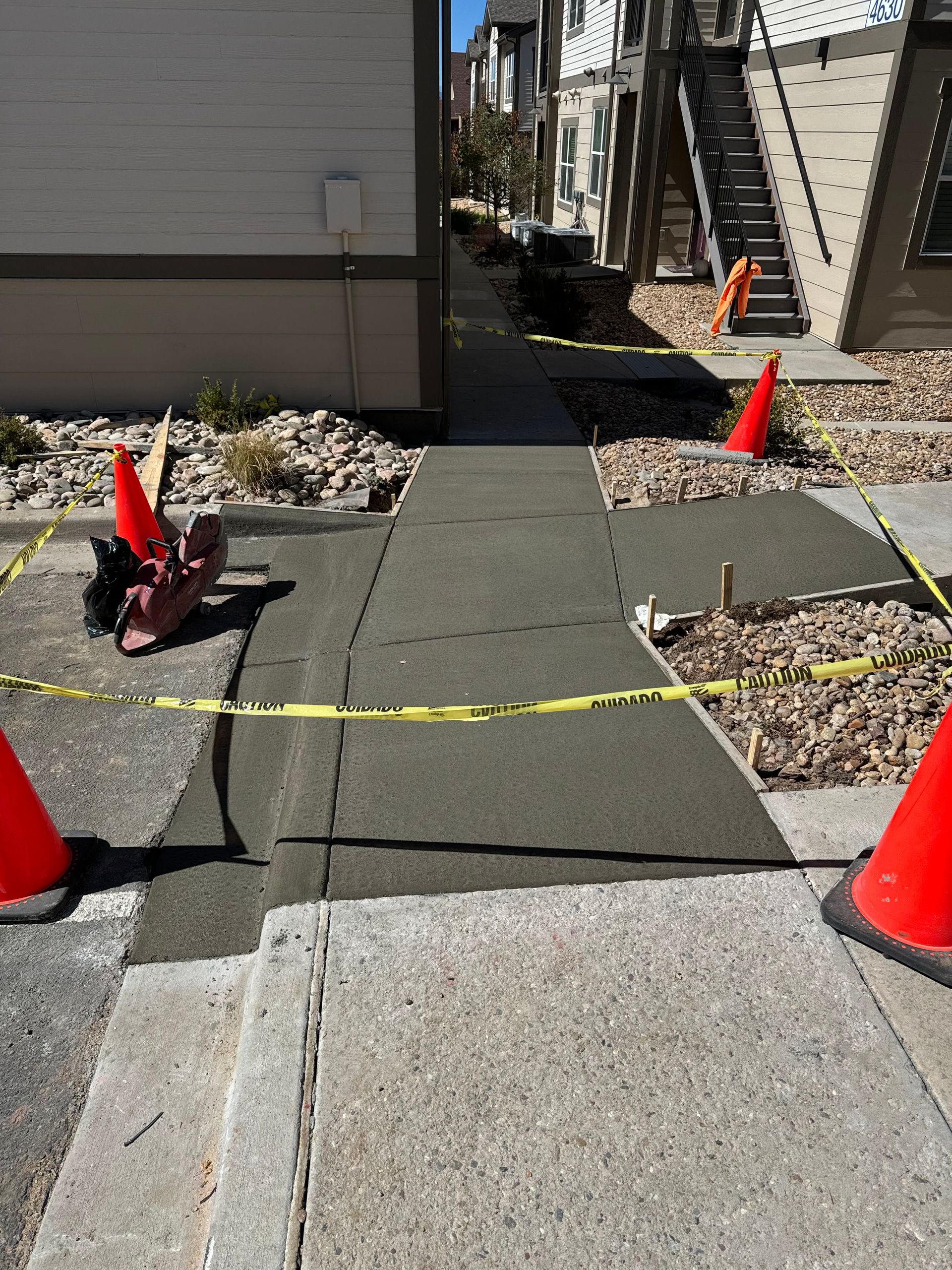 Newly poured concrete sidewalk section with caution tape and cones in an urban setting. The walkway is between buildings.