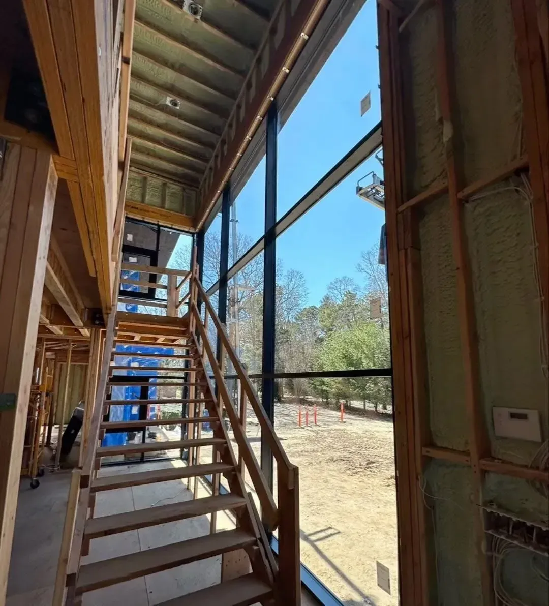 Interior of a building under construction, featuring a wooden staircase and large windows with a view.