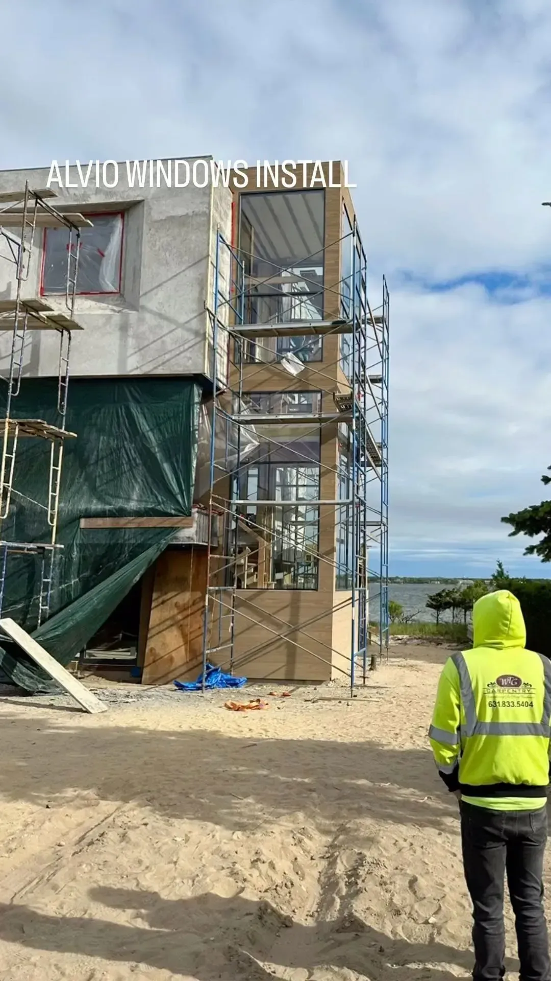 Construction worker in neon jacket, viewing building with scaffolding on a sandy beach.