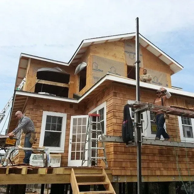Construction workers on a two-story house, sawing and working on the exterior under a cloudy sky.