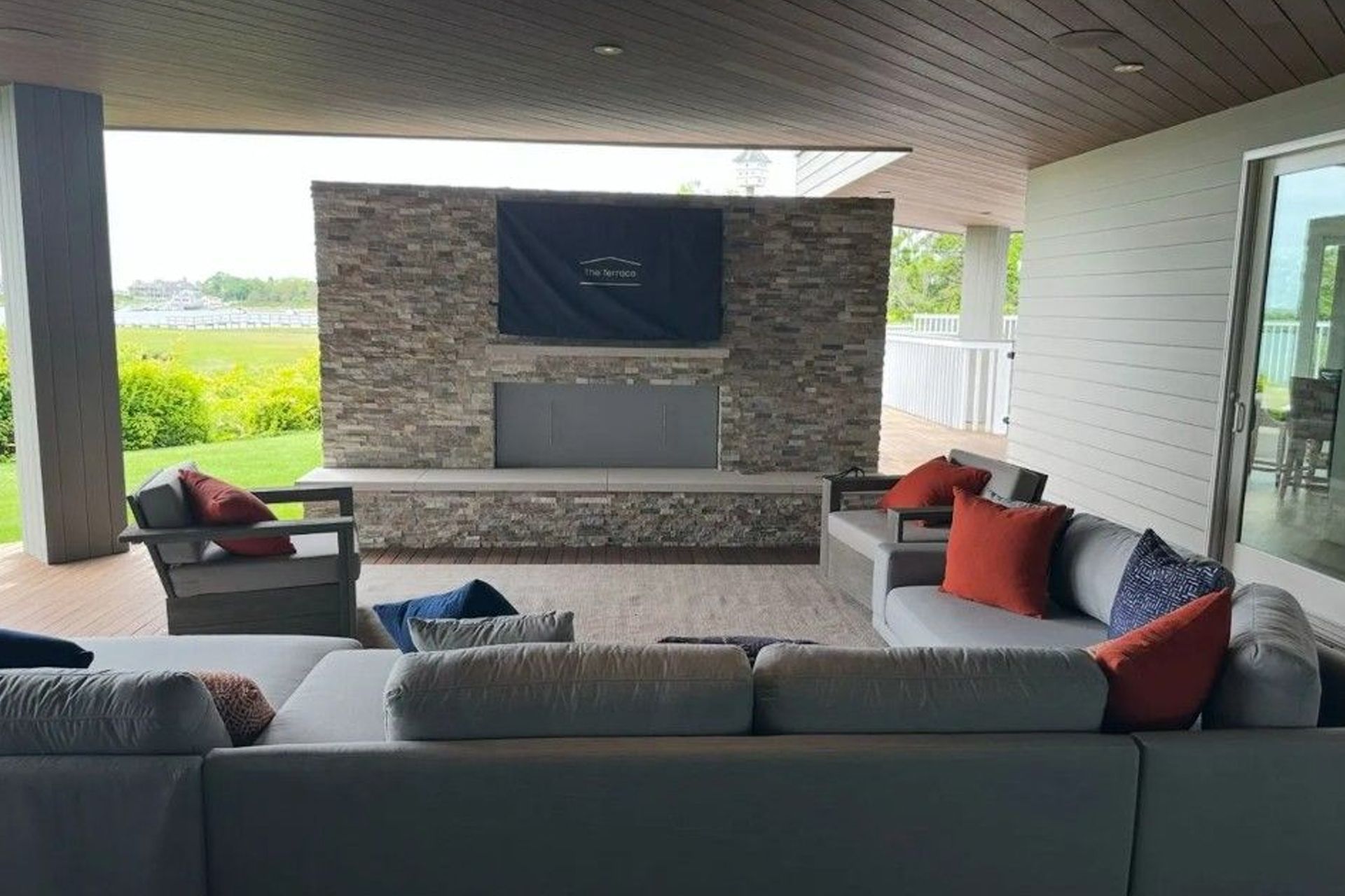 Covered outdoor TV above a stone fireplace. Gray sectional sofa with orange and blue cushions on a wooden patio.