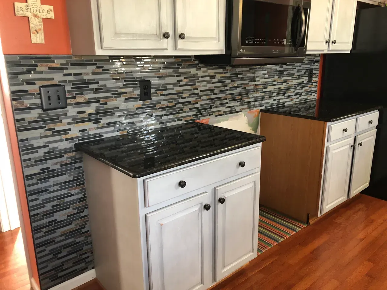 A kitchen with white cabinets , black counter tops , a microwave and a cross on the wall.
