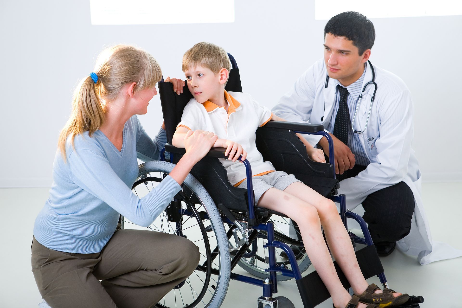 Woman and doctor with child in wheelchair; all looking at the child in a bright room.