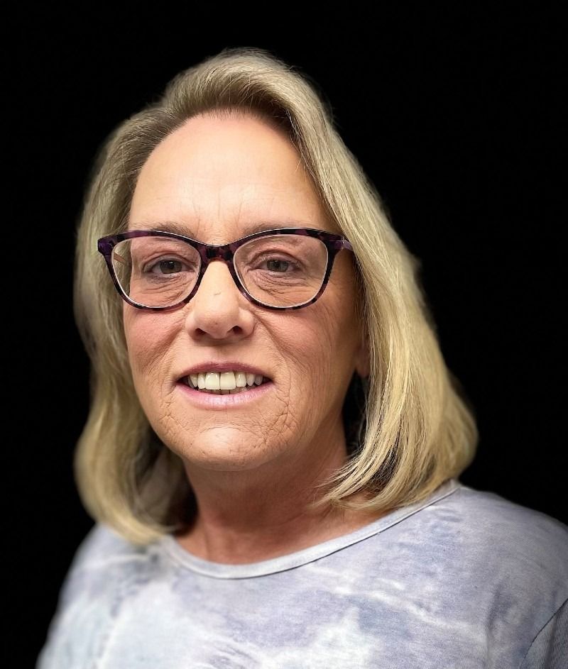 Woman with blonde hair, wearing glasses and a grey tie-dye shirt, smiling at the camera.