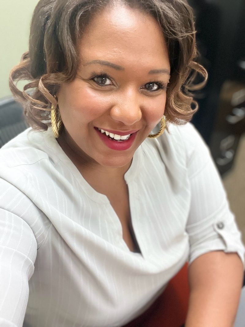 Woman with curly brown hair, smiling, wearing gold earrings and a light gray blouse.