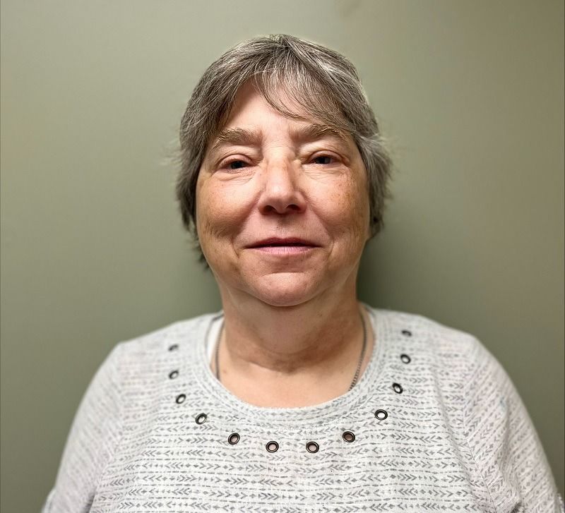 Woman with short gray hair and a gray patterned sweater smiles in front of a green wall.