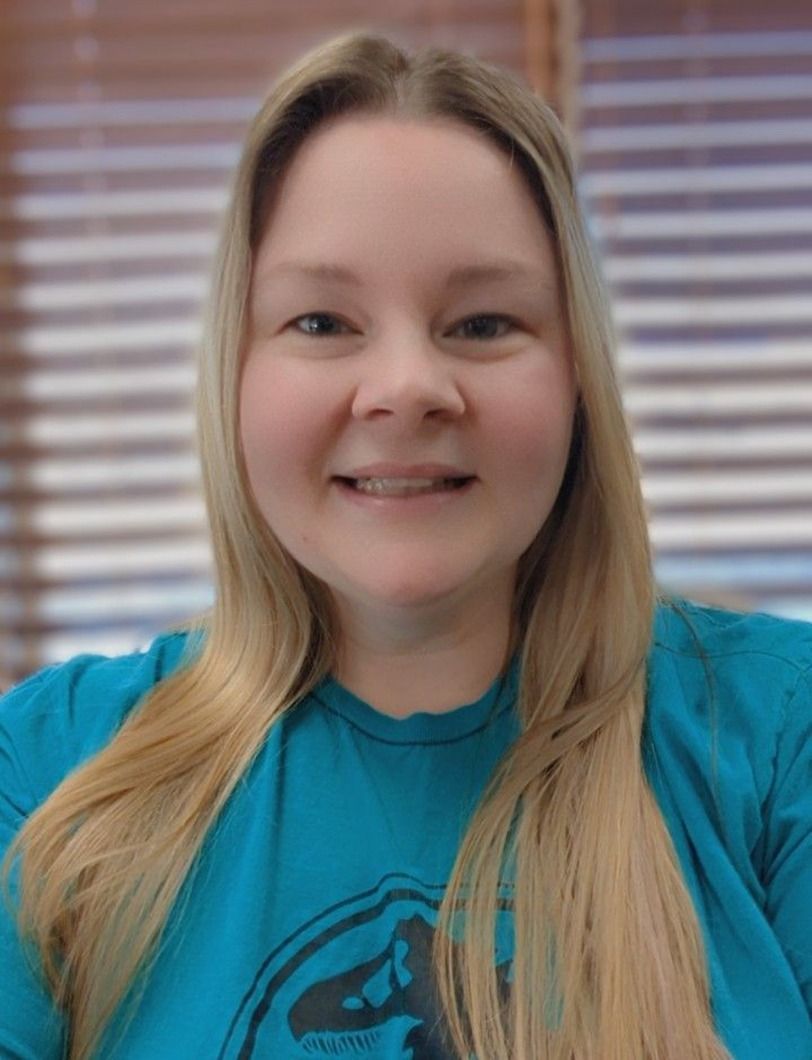Woman with blonde hair smiling, wearing a teal t-shirt, in front of a window with blinds.