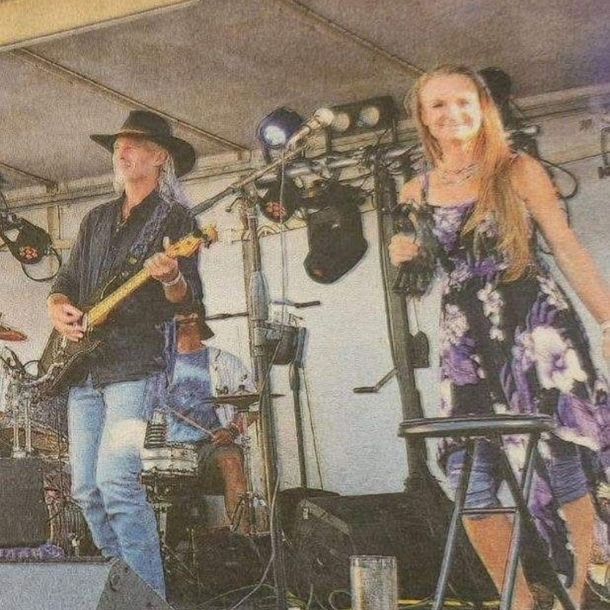 A band performs on stage; a guitarist in a cowboy hat plays while a singer in a floral dress sings with a microphone.