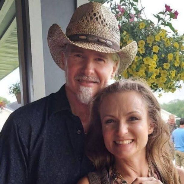 A man in a cowboy hat and a woman smiling, outdoors by flowers.