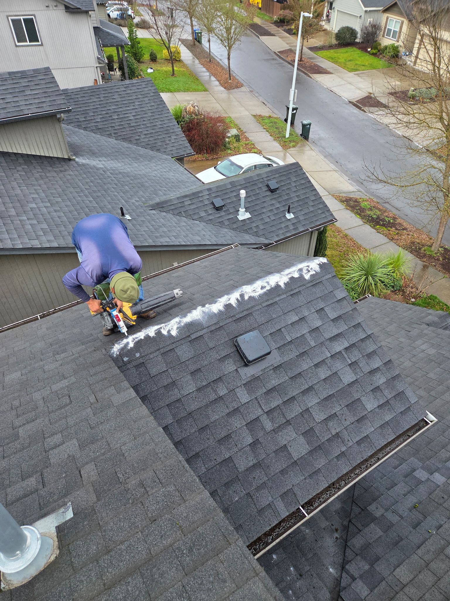 Person on a dark shingled roof, cleaning gutters. Overcast day, residential street visible.