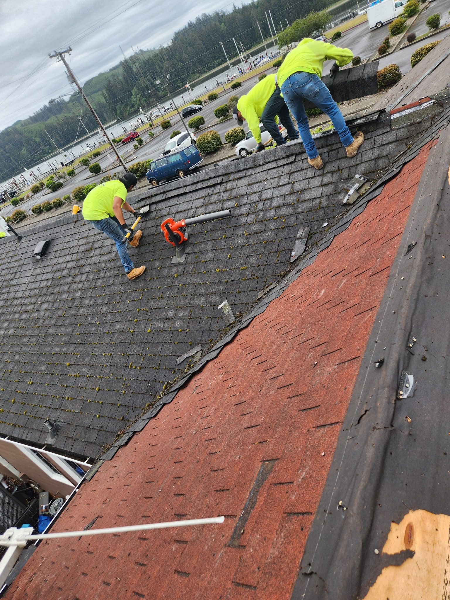 Roofers in neon vests work on a dark shingle roof, cloudy sky.