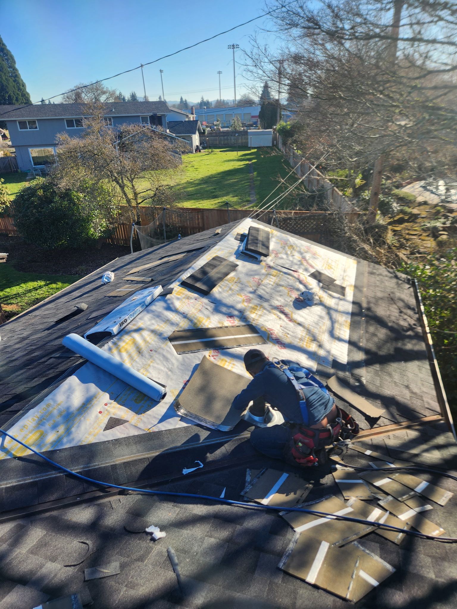 Roofer on a roof, removing old shingles. Sunny day, residential setting with houses and trees.