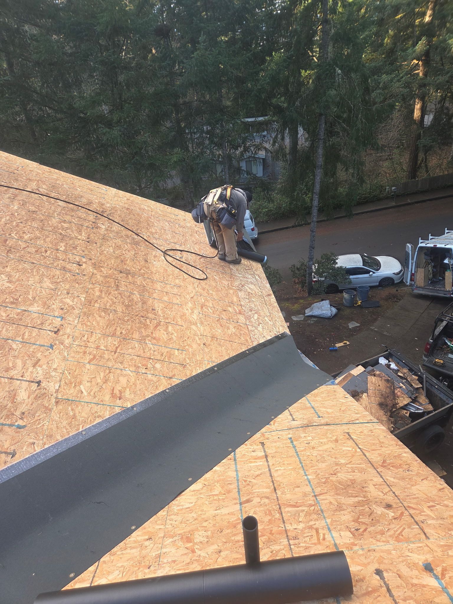 Roofer on a roof installing flashing. Brown wood, black flashing. Trees and road in background.