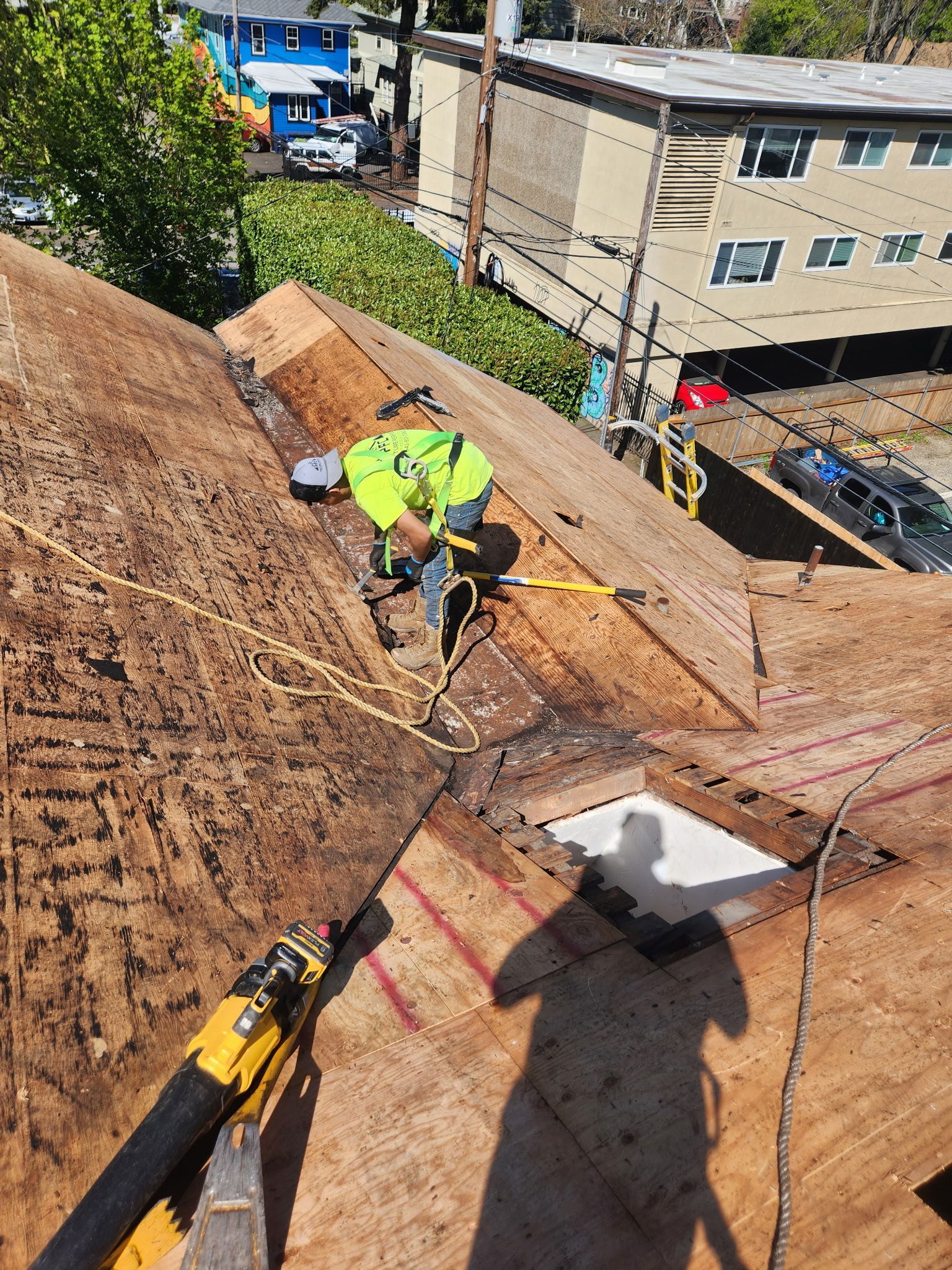 Roofer in safety gear works on a rooftop with plywood, tools, and city buildings in the background.