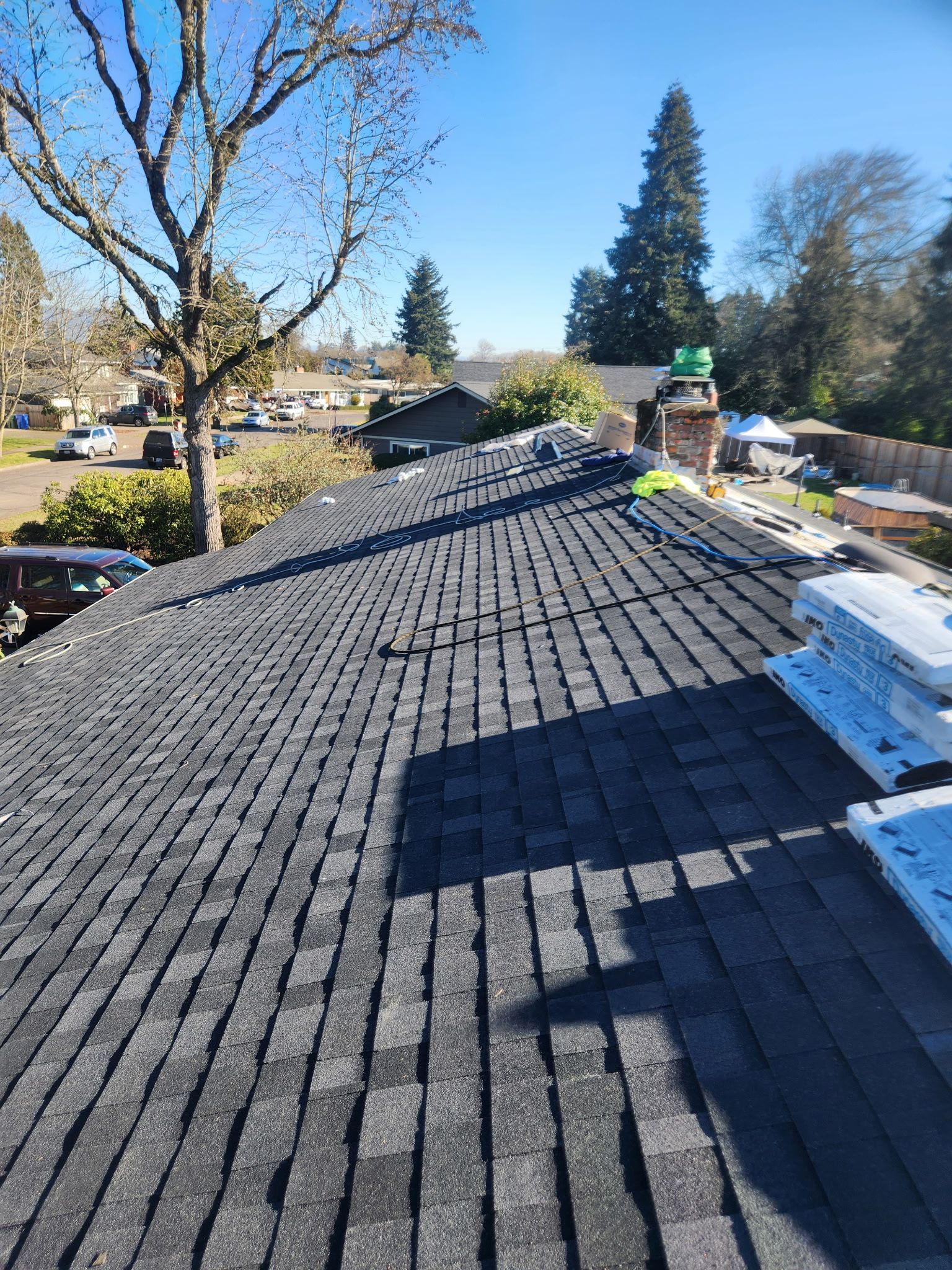 Dark asphalt shingle roof on a house, partially completed, under a bright blue sky.