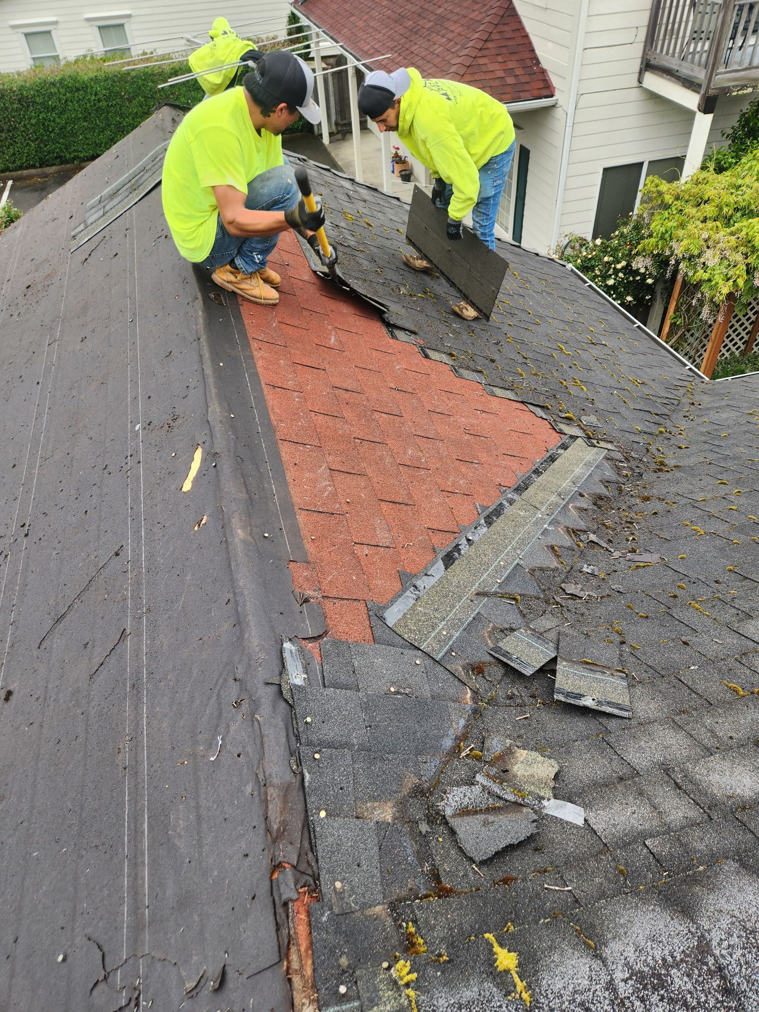 Two roofers in neon vests repair a damaged roof, one using a tool, the other holding a shingle.
