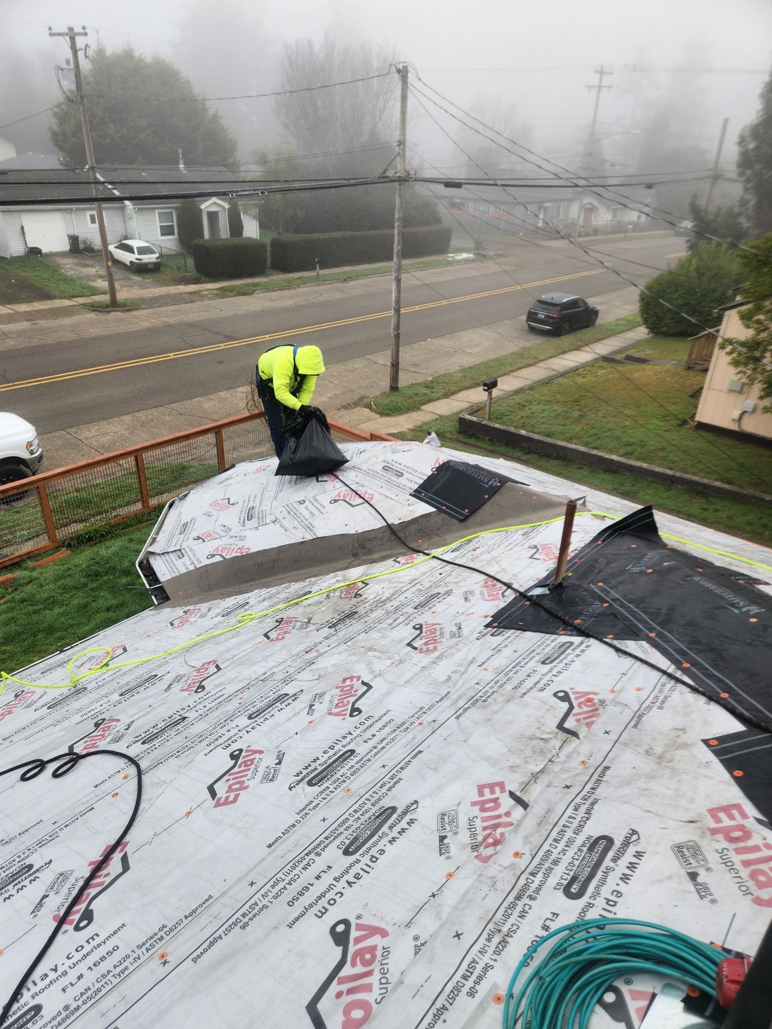 Roofer in safety vest on a roof, foggy day. Collecting debris. Residential street in the background.