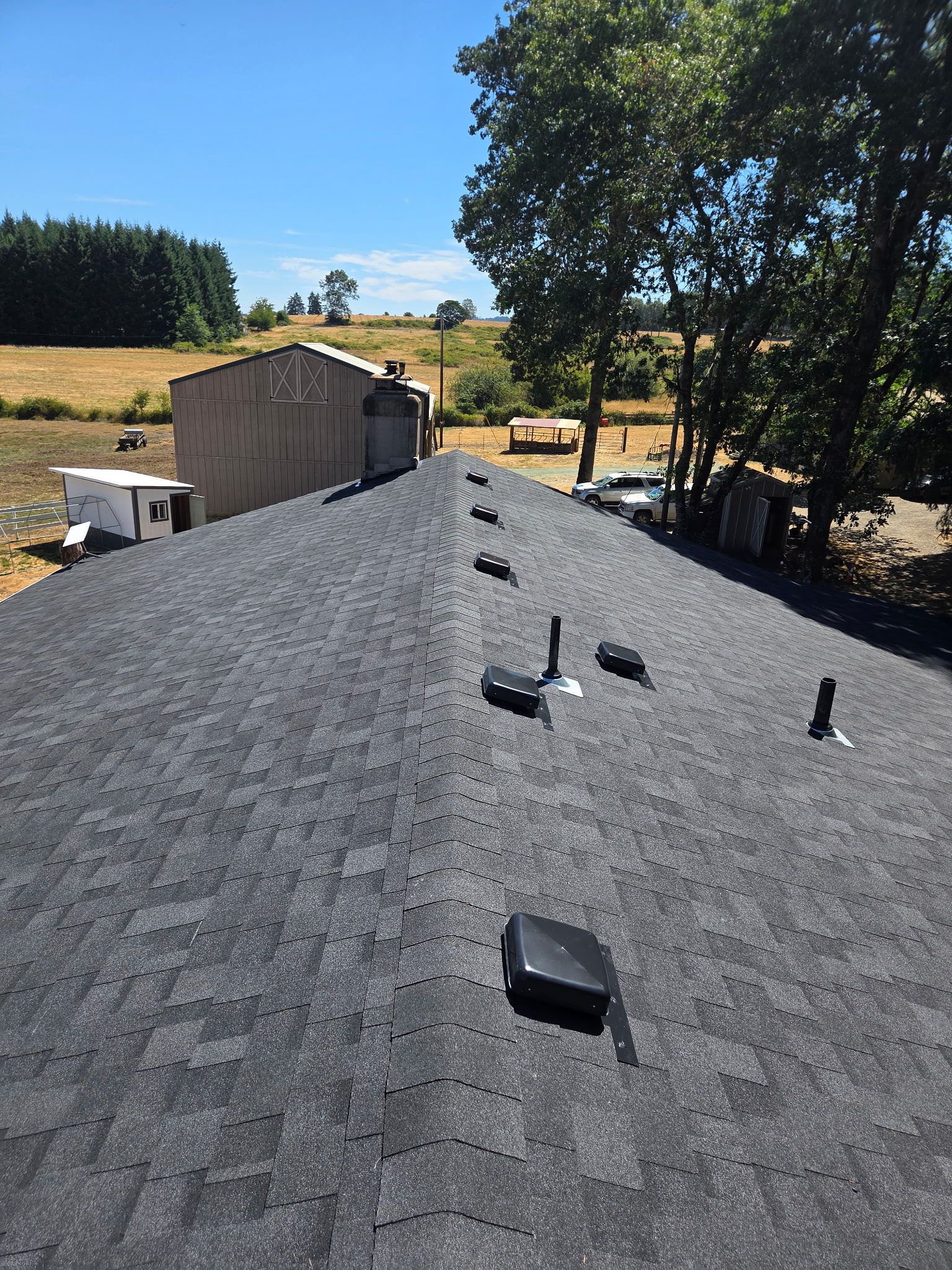 View from a rooftop with dark shingles, overlooking a rural landscape with buildings and trees under a blue sky.