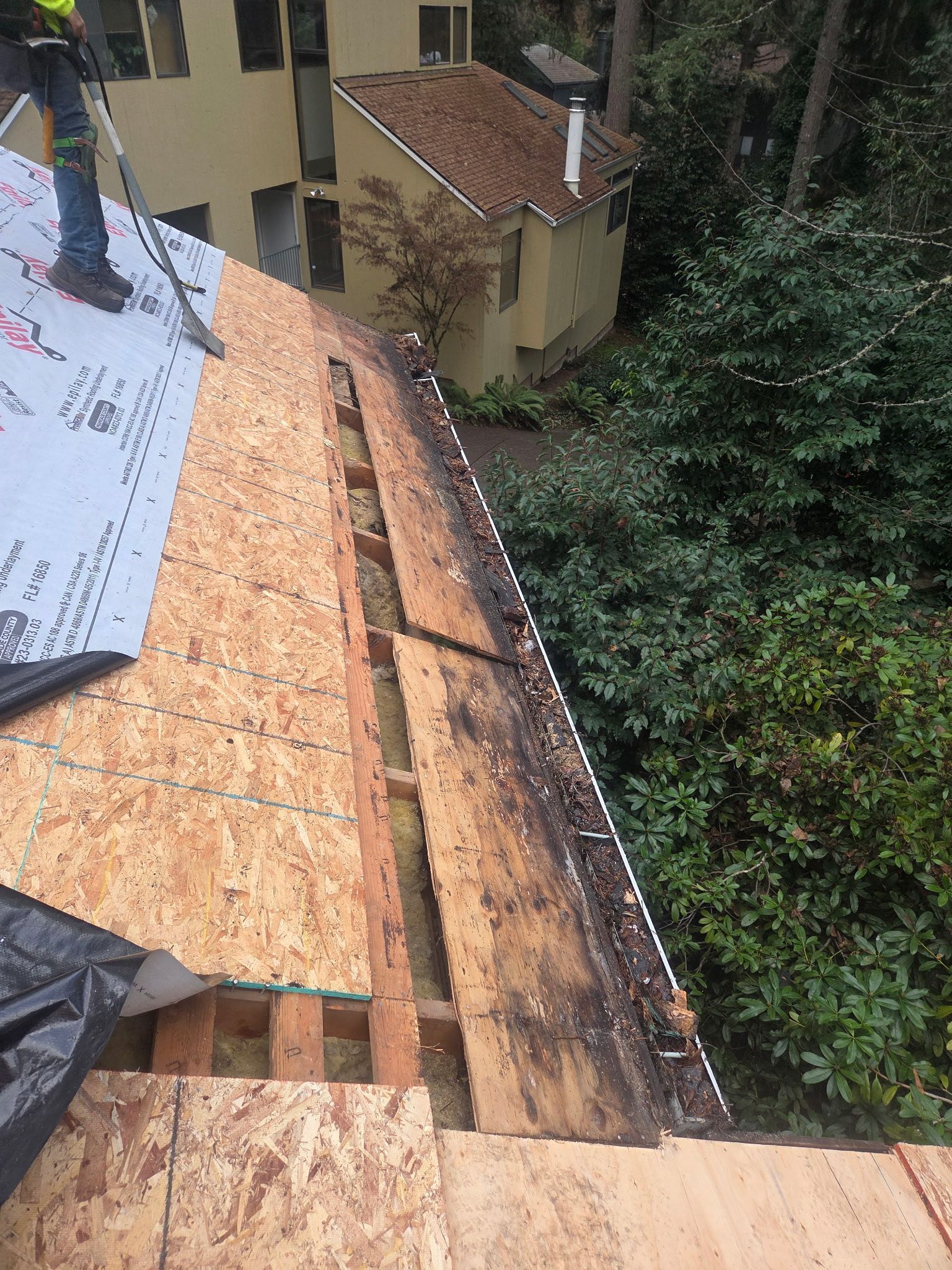 Roofer on a partially built roof, applying underlayment. Wood, dark staining, and trees visible.