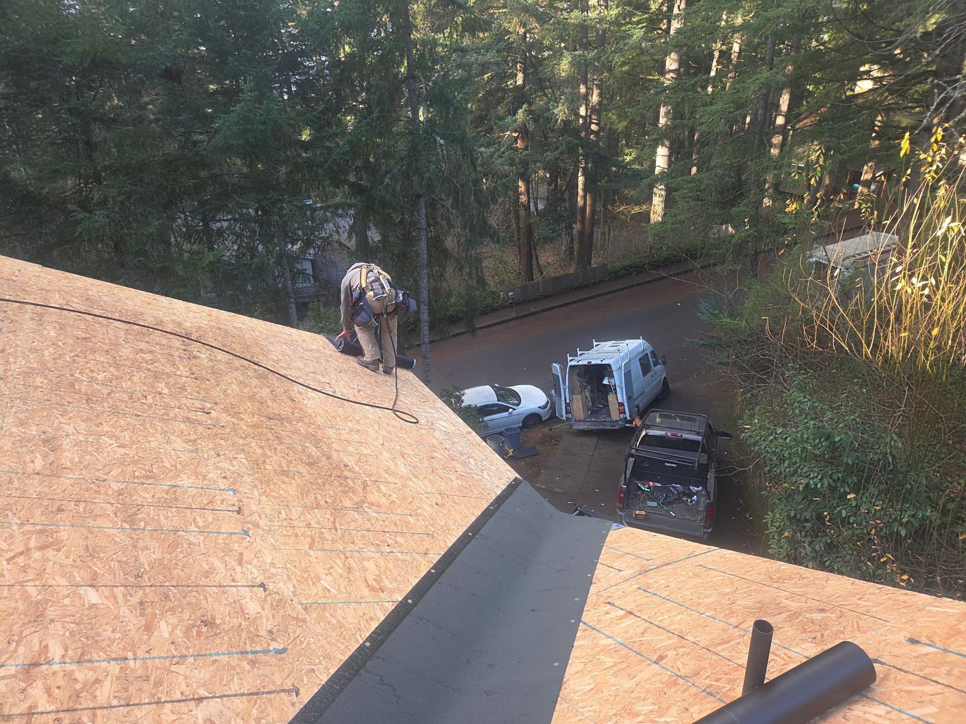 Roofer on a partially shingled roof, working near a black gutter. Cars and trees are in the background.