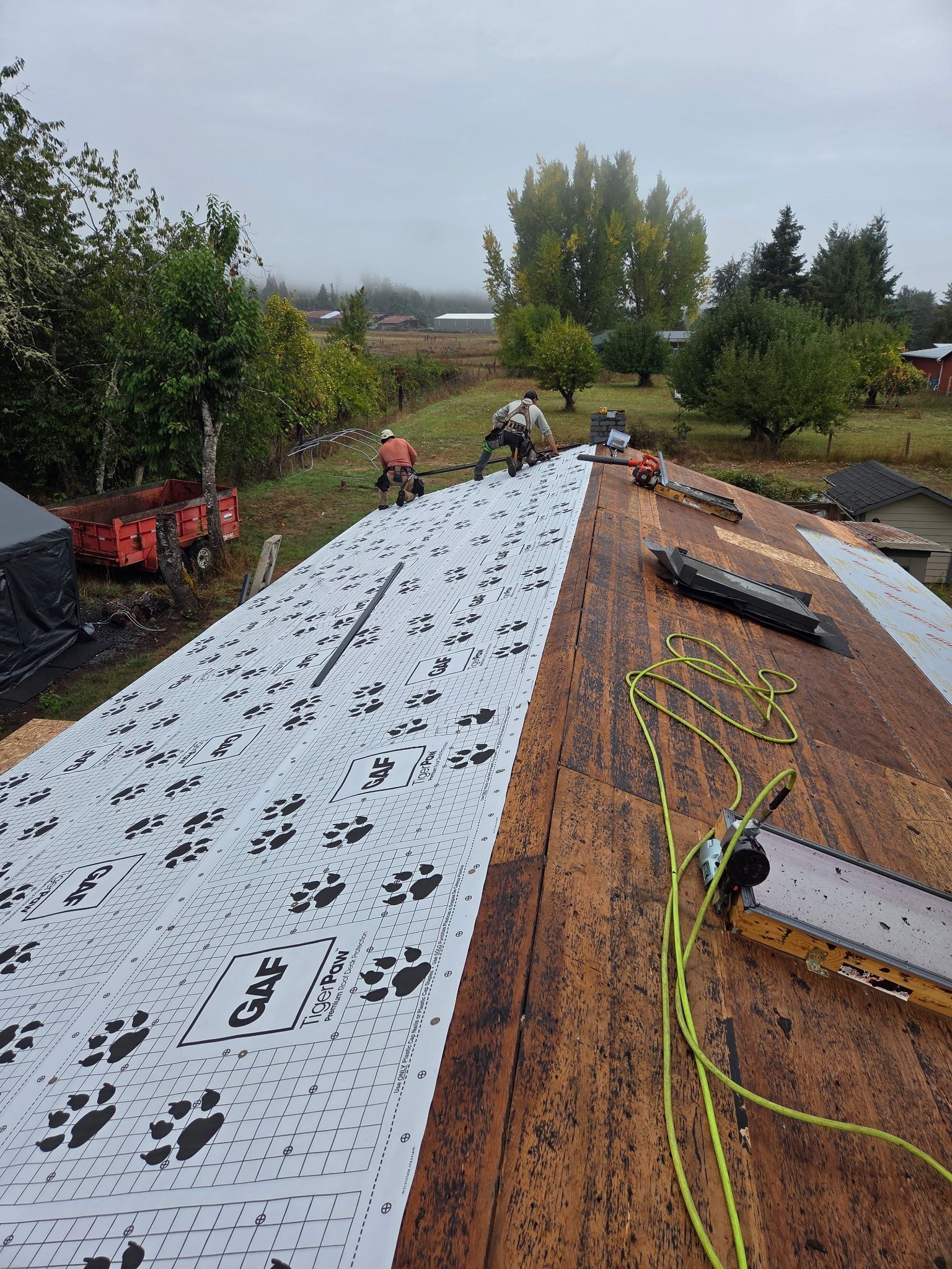 Roofers installing underlayment on a house roof. Landscape and tools visible. Overcast day.