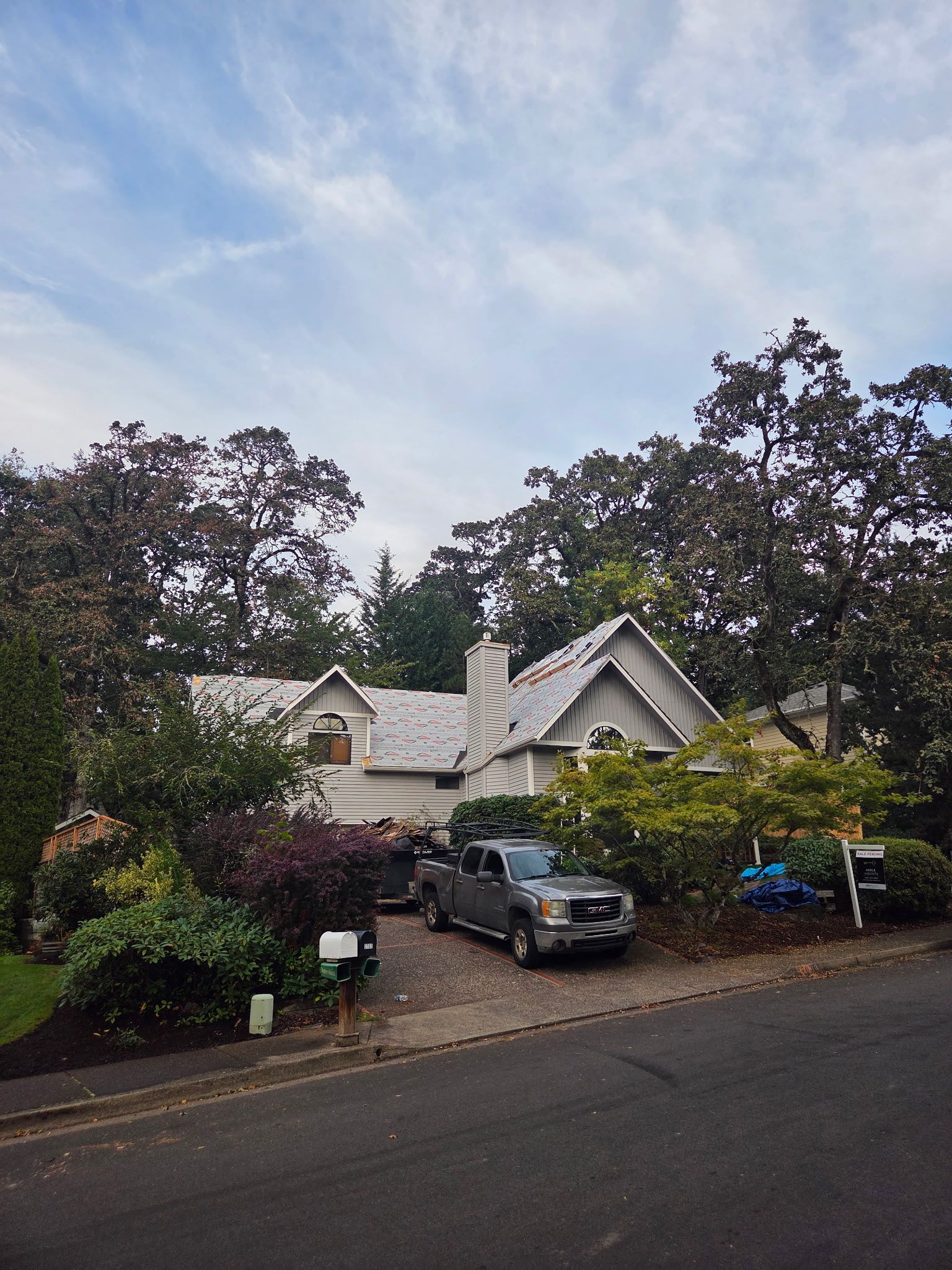 House with white siding and gray roof, surrounded by trees and shrubbery, truck parked in driveway.