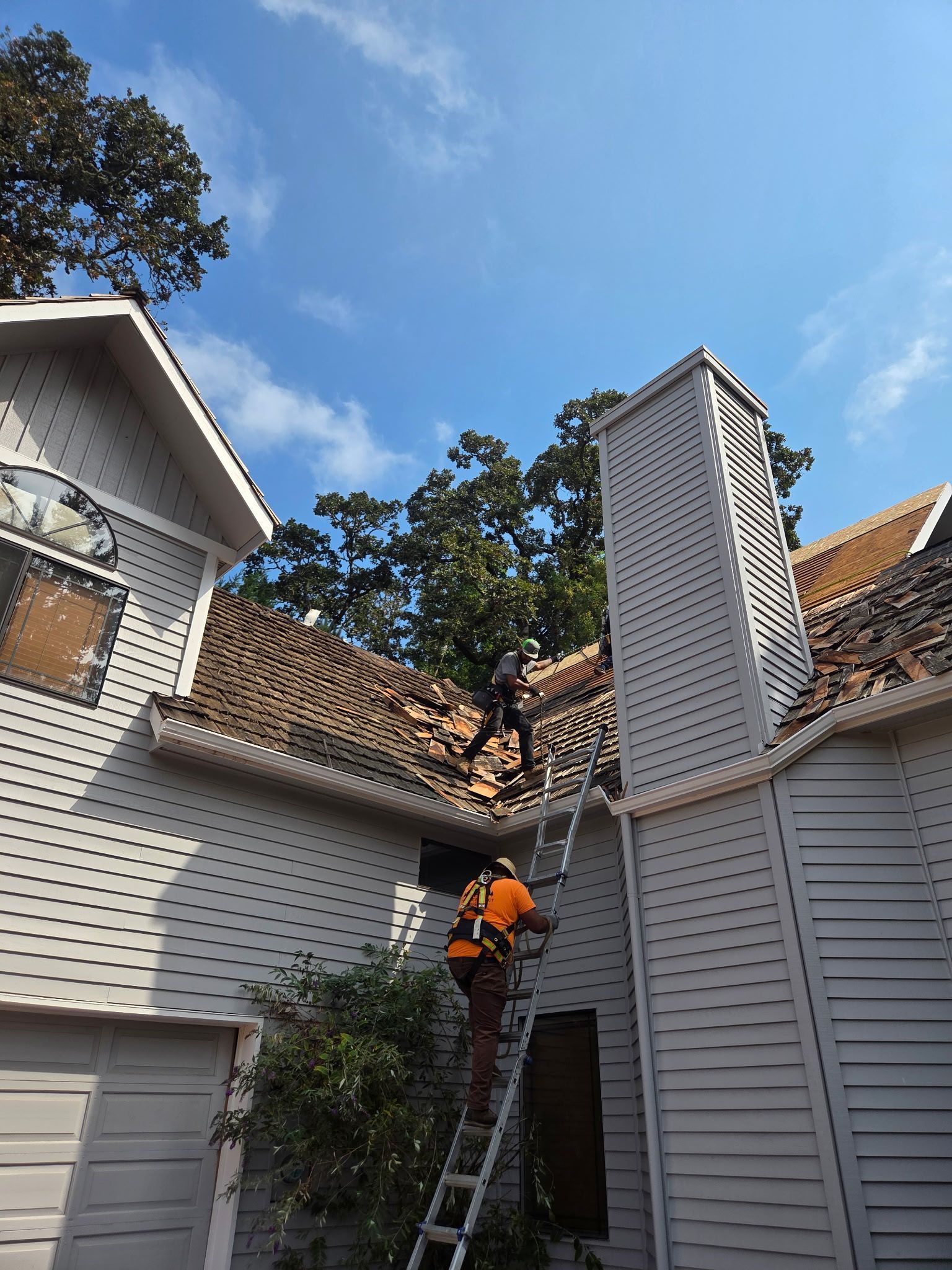 Person on ladder, working on roof with damaged shingles; blue sky, chimney, and gray house visible.
