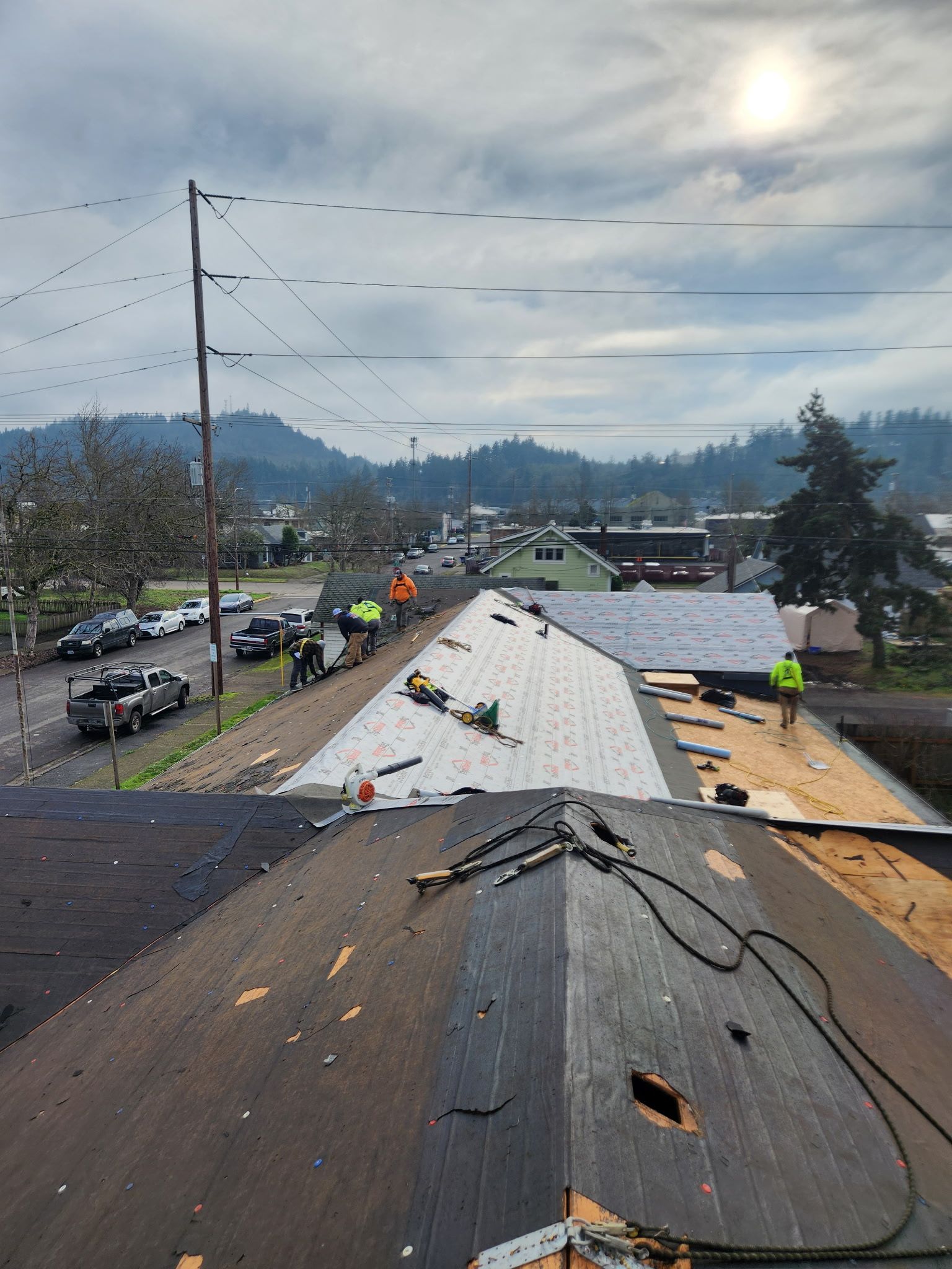 Workers repairing a roof with a view of a street lined with cars and buildings under a cloudy sky.