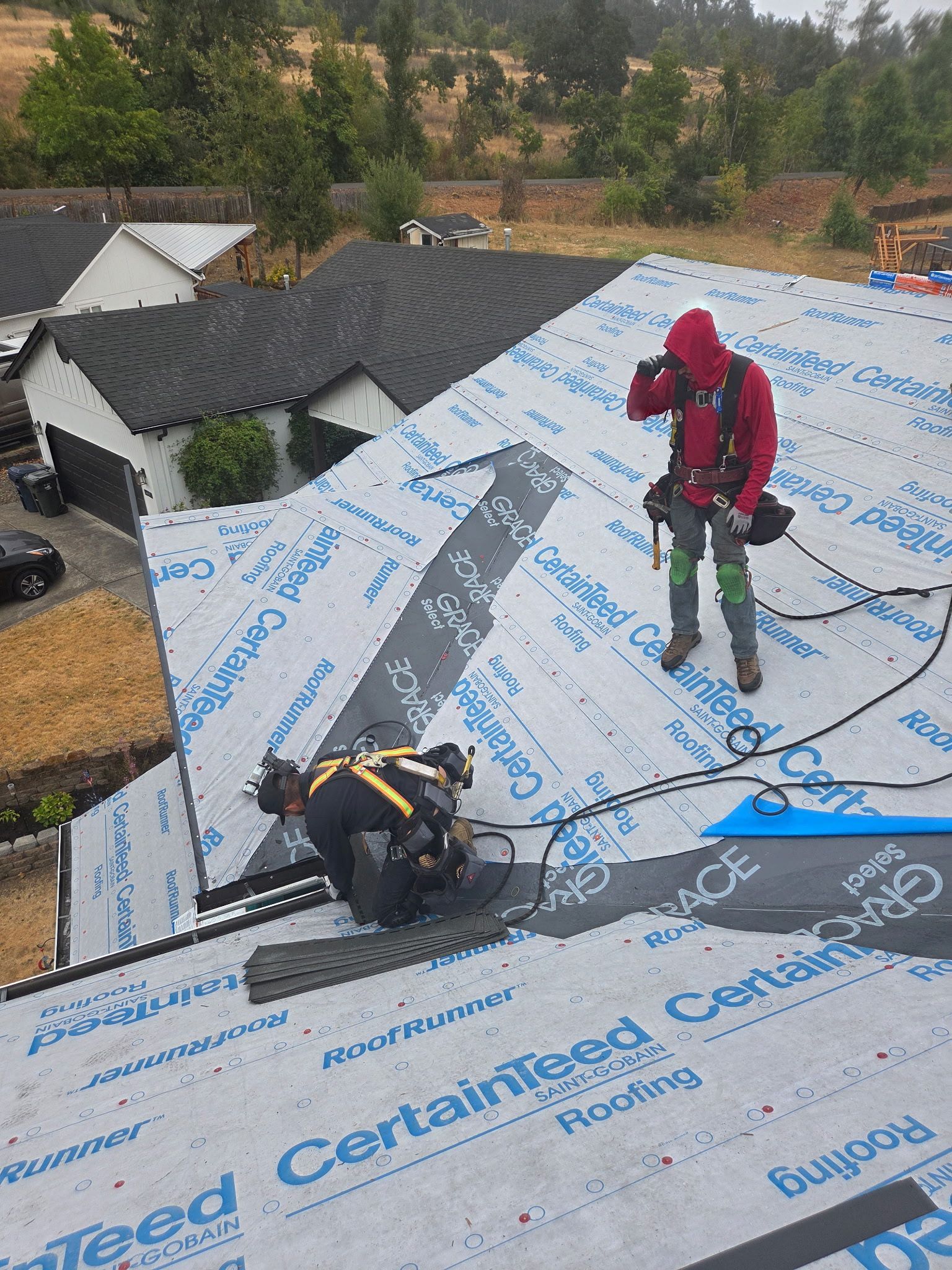 Two roofers working on a house roof, one cutting, the other standing; black shingles, blue underlayment.