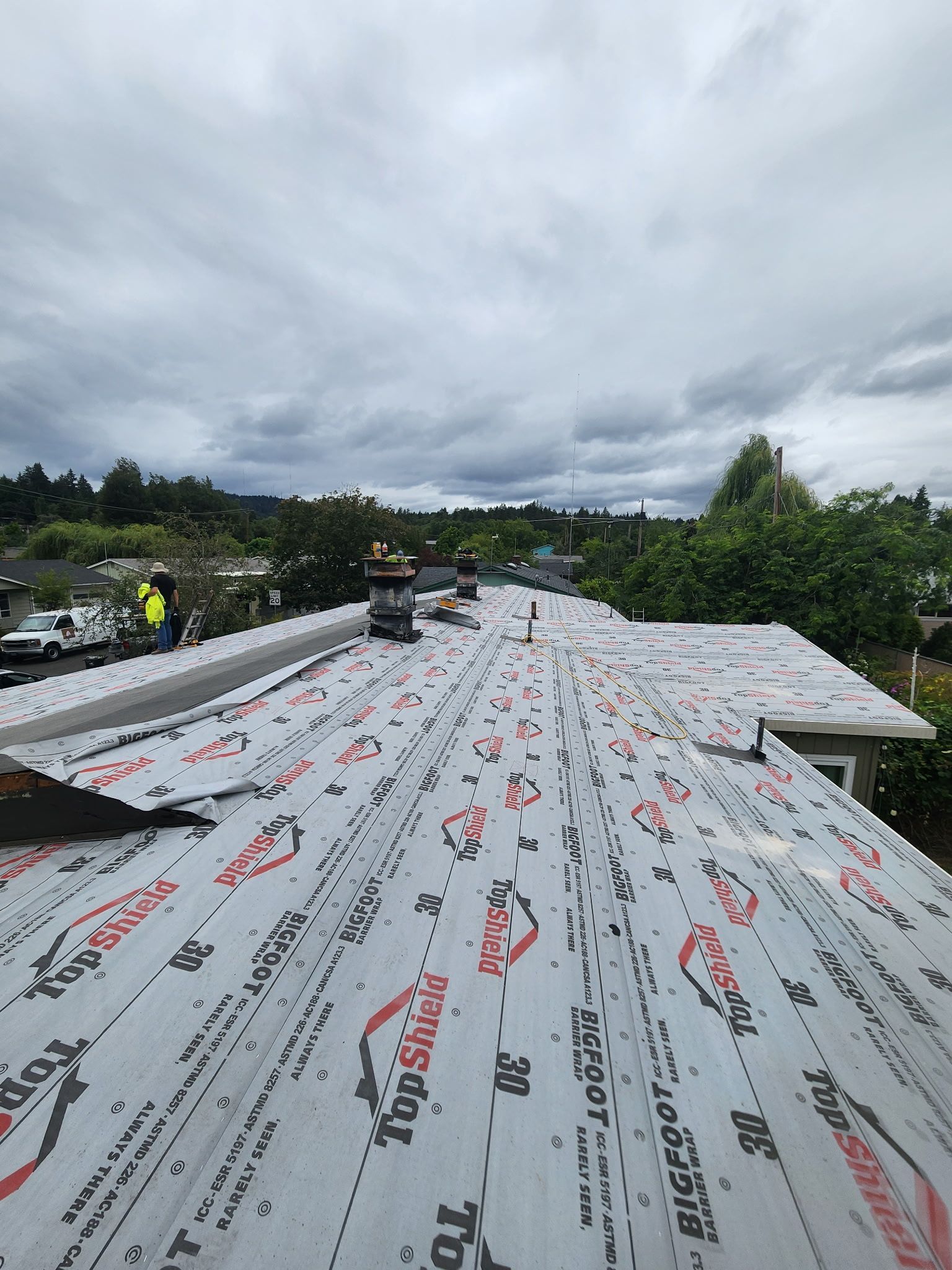 Rooftop with white underlayment, several chimneys, and a cloudy sky. Trees and houses in the background.