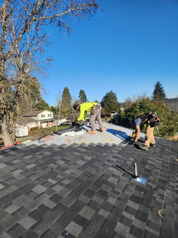 Two roofers installing shingles on a roof under a blue sky, surrounded by trees and houses.