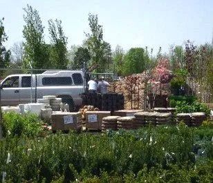 Truck loading gardening supplies at a nursery. Two workers, green plants, brown boxes, and a sunny day.