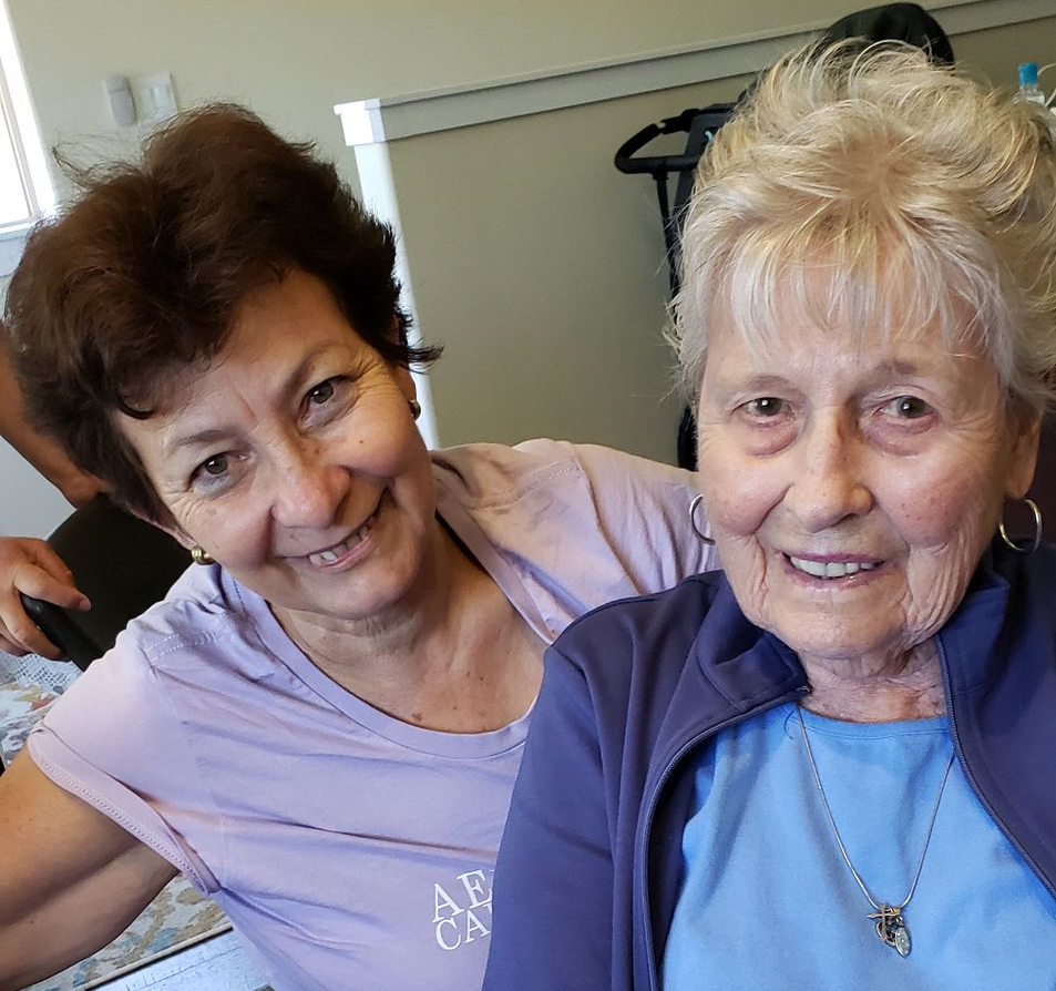 Woman embracing and smiling at an older woman seated on a couch indoors.