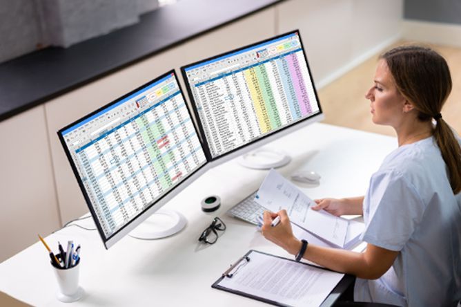 Woman in scrubs reviews documents at a desk with two monitors displaying spreadsheet data