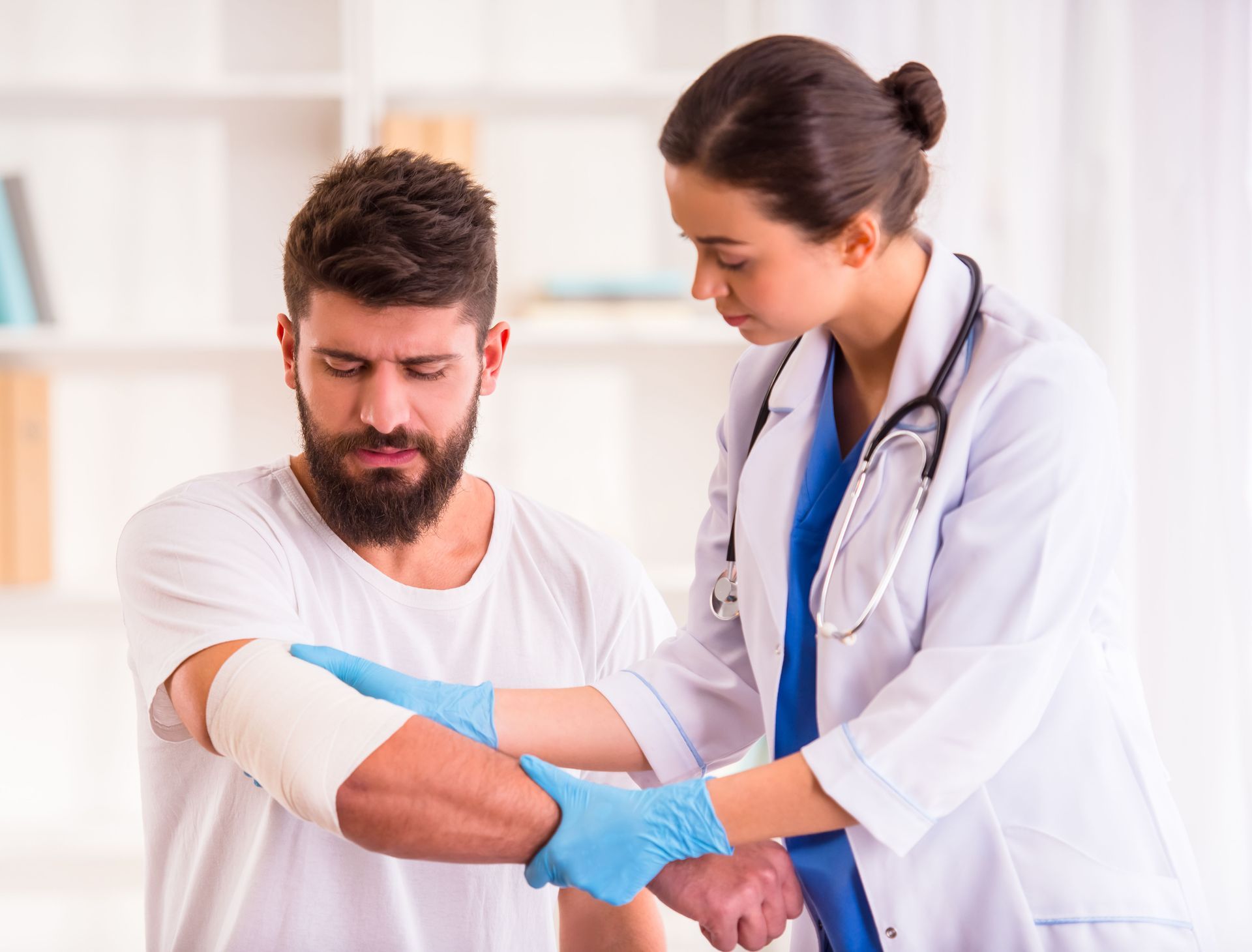 A doctor examines a man's bandaged arm. Doctor wears gloves and a stethoscope.