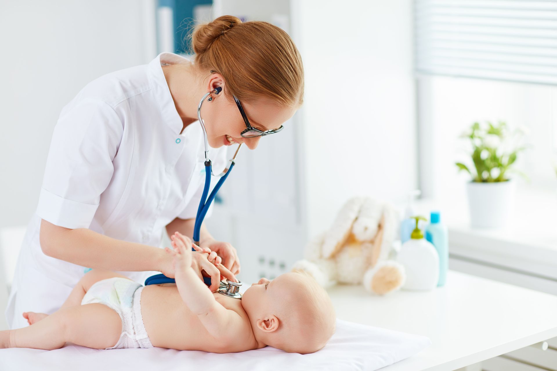 Doctor listens to baby's heartbeat with stethoscope in a bright room. The baby lies on a white towel on a table.